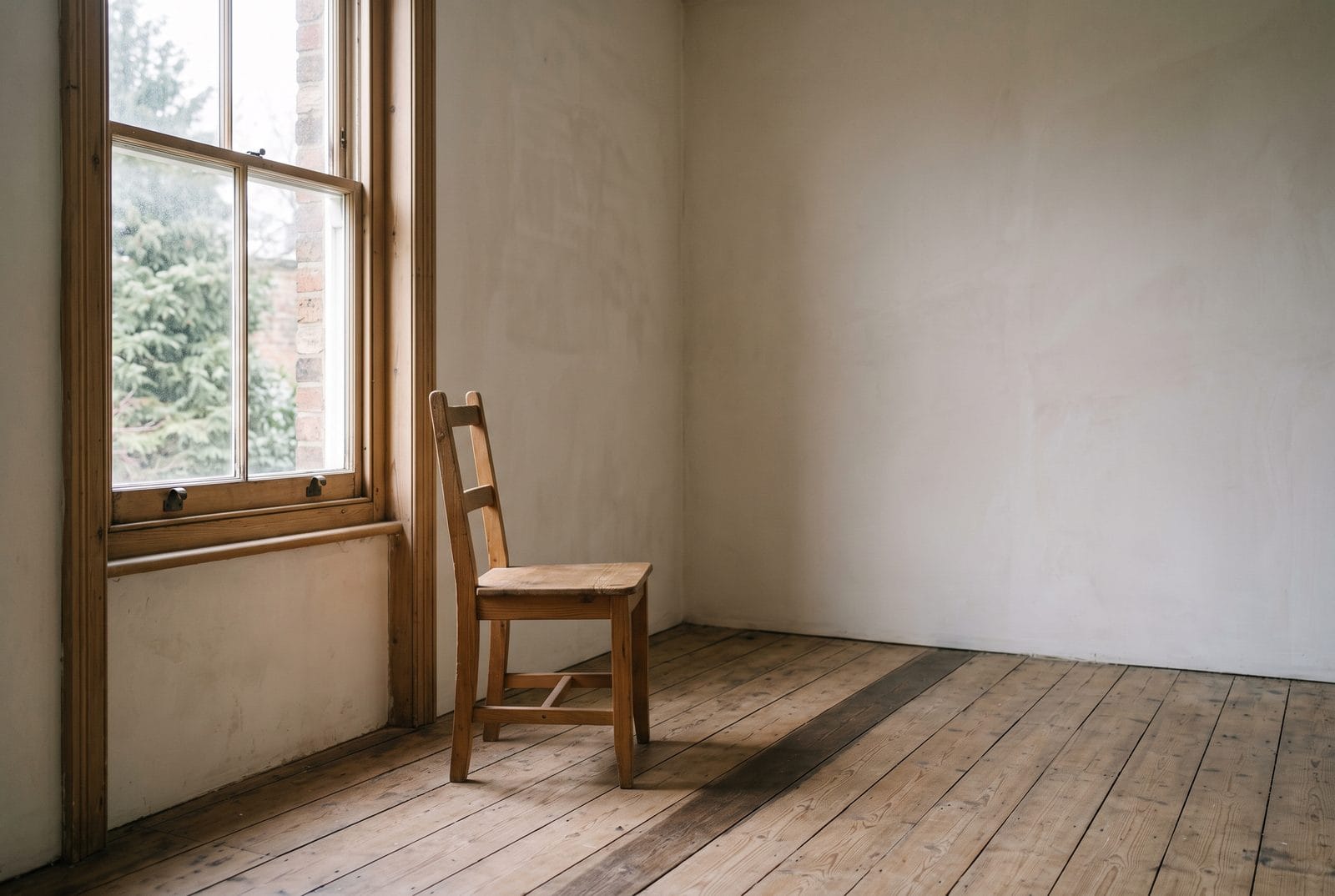 An empty room with one wooden chair beside a tall window and bare floorboards